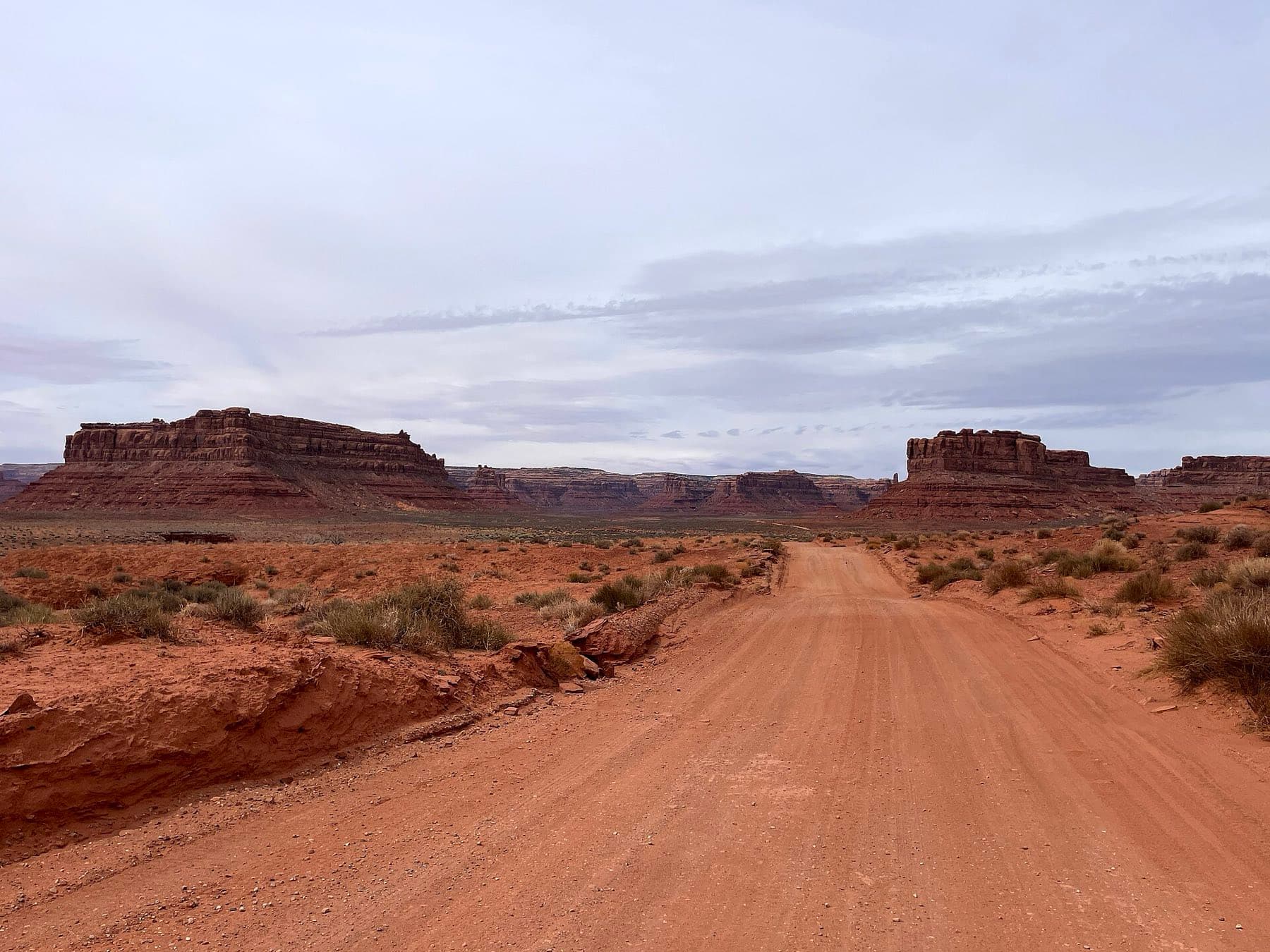 A red dirt road running toward flat-topped sandstone mesas under a wide overcast sky in the Four Corners high desert